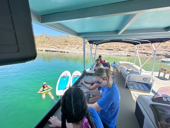 Group relaxing on a shaded pontoon boat over emerald-green lake water with two paddleboards and a swimmer near a rocky, cactus-studded desert shoreline on a sunny day.