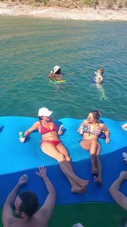 Two women in bikinis lounging with drinks on a large blue floating mat at a sunny lake, swimmers nearby in clear water and a rocky shoreline in the background