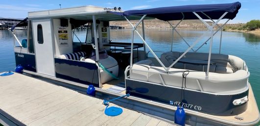 Blue-and-white pontoon houseboat with canopy and cushioned seating docked at a calm lake marina on a sunny day