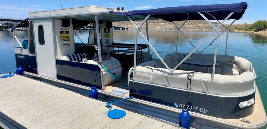 Blue-and-white pontoon houseboat with canopy and cushioned seating docked at a calm lake marina on a sunny day