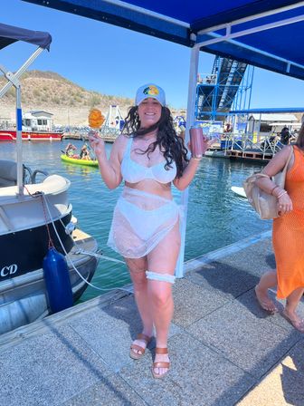 Woman in white bikini and sheer wrap on a sunny marina dock wearing a baseball cap, holding a spiral potato snack on a stick and a tumbler, smiling with boats, kayakers and blue dock canopy in the background.