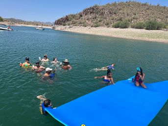 Sunny desert lake scene with a group of people swimming and relaxing on a bright blue floating mat, some holding drinks, near a cactus-covered shoreline and boats in the distance.