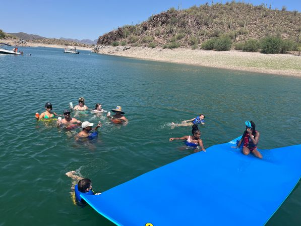 Sunny desert lake scene with a group of people swimming and relaxing on a bright blue floating mat, some holding drinks, near a cactus-covered shoreline and boats in the distance.