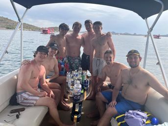 Eight friends in swim trunks posing on a pontoon boat on a calm lake, drinks on the center table and rocky desert shoreline in the background