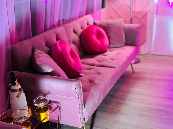Pink velvet tufted sofa in a neon-lit lounge with bright round pink pillows, a champagne bottle and coupe glass on a glass side table, and light wood flooring.