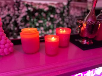 Three glowing red candles on a pink-lit mantel, floral wall backdrop and a metallic champagne bucket nearby