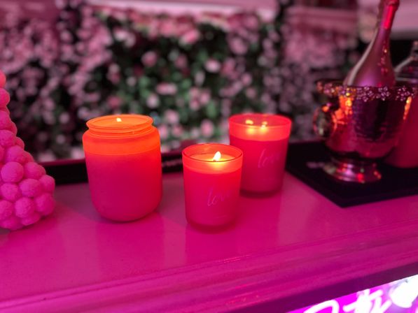 Three glowing red candles on a pink-lit mantel, floral wall backdrop and a metallic champagne bucket nearby