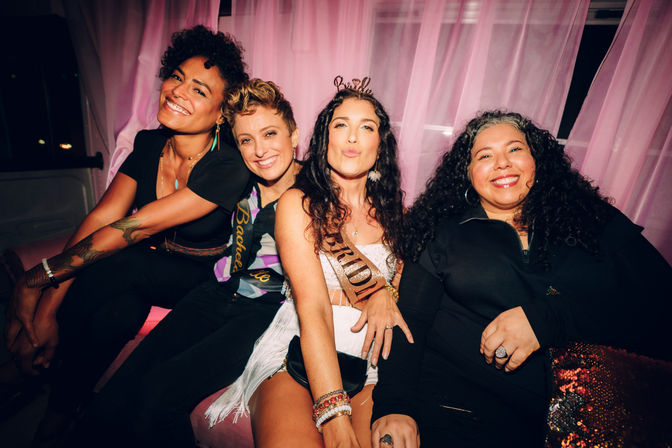 Four friends on a pink-curtained couch at an indoor bachelorette party, bride-to-be wearing a sash and tiara making a kiss face while the others smile