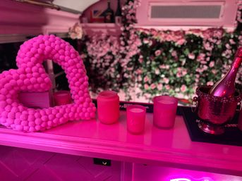 Valentine-themed pink-lit display on a counter with a fluffy pom-pom heart wreath, three 'love' candles, and a metallic ice bucket holding a champagne bottle in front of a rose-covered floral wall.