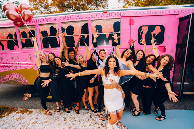 Bachelorette party: bride in white fringe dress and veil with rose-gold balloons, surrounded by friends in black dresses cheering with hands raised in front of a bright pink party trolley