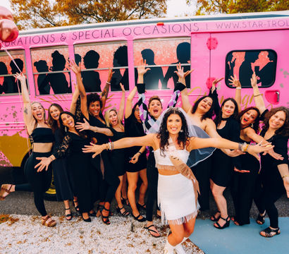 Bachelorette party: bride in white fringe dress and veil with rose-gold balloons, surrounded by friends in black dresses cheering with hands raised in front of a bright pink party trolley