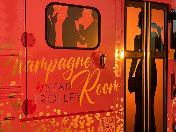 Bright coral-orange trolley side with gold script reading "Champagne Room", confetti dots and window silhouettes of people holding drinks backlit by warm sunset light.