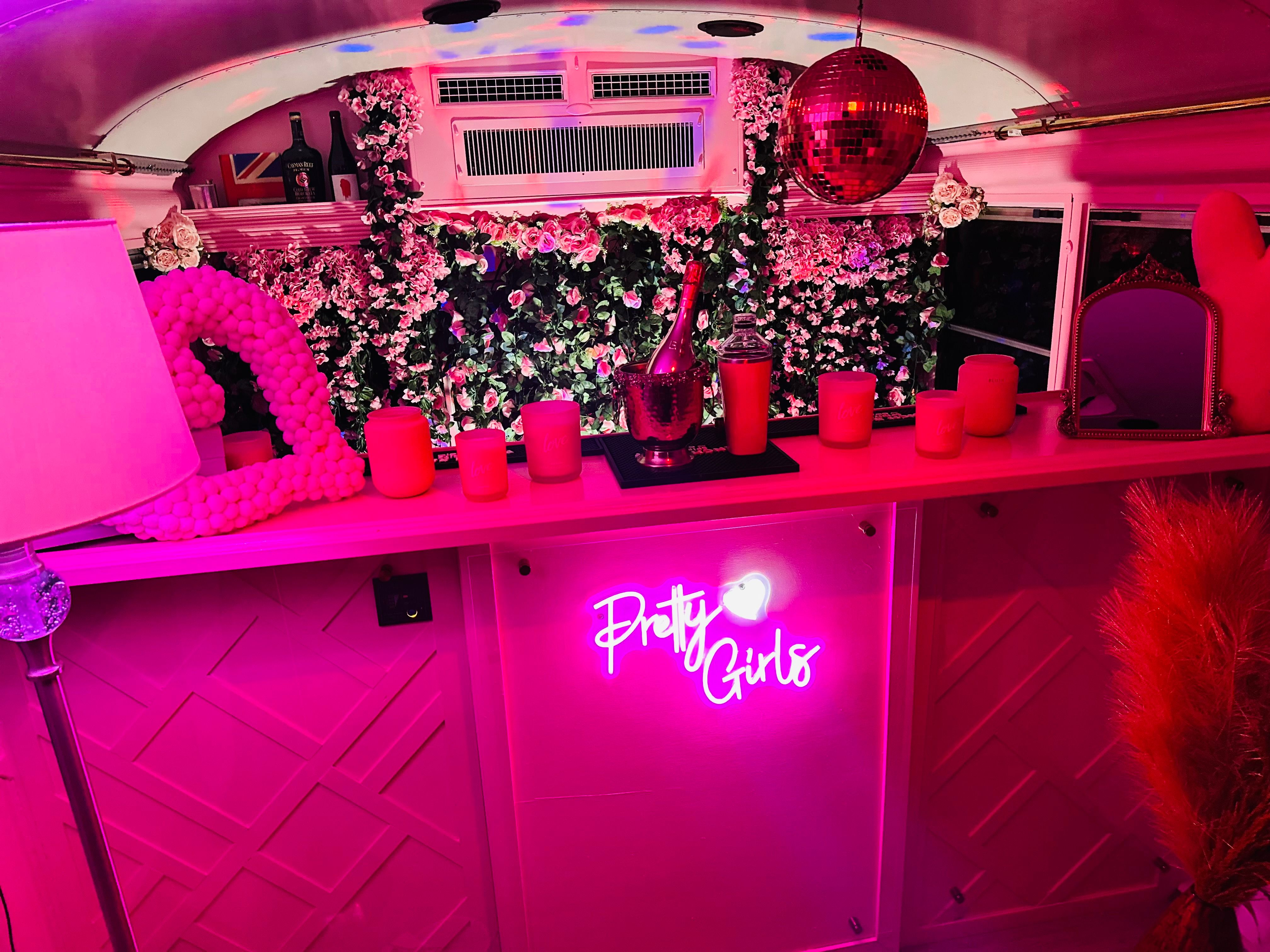 Vibrant pink party bus interior with a flower wall backdrop, disco ball, champagne bucket, candles and a glowing 'Pretty Girls' neon sign — playful party decor.