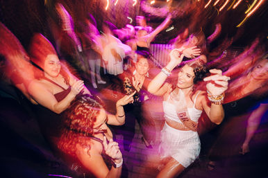 Energetic bachelorette party dancing on a nighttime patio with colorful motion-blur lights and lively crowd