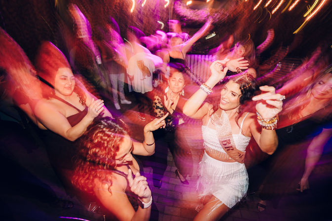 Energetic bachelorette party dancing on a nighttime patio with colorful motion-blur lights and lively crowd