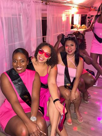 Group of women in pink dresses with black sashes posing in a pink-lit party lounge, smiling and wearing playful sunglasses.