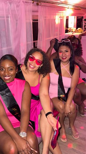 Group of women in pink dresses with black sashes posing in a pink-lit party lounge, smiling and wearing playful sunglasses.
