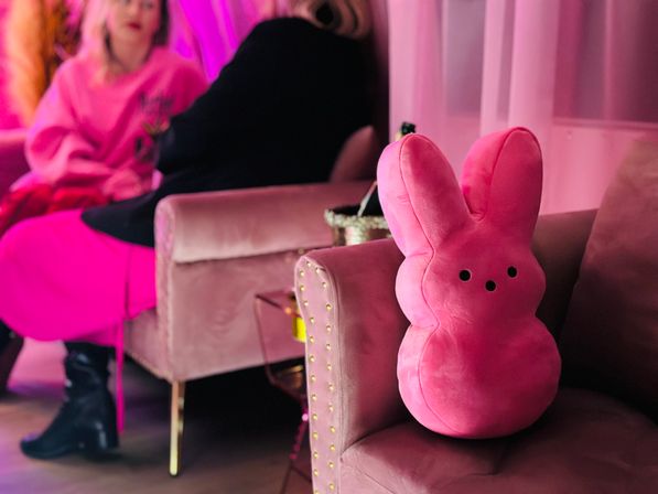 Plush pink bunny pillow perched on a studded velvet armchair in a pastel-pink lounge, with blurred figures and cozy decor in the background.