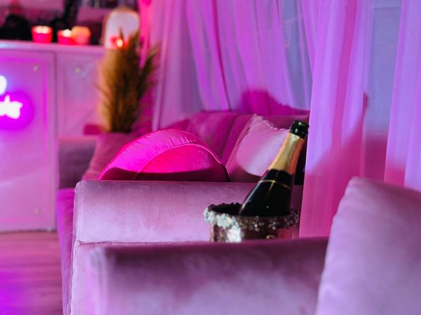 Pink velvet lounge seating with plush pillows, a chilled champagne bottle in an ice bucket, sheer curtains and neon ambient lighting creating a cozy VIP vibe