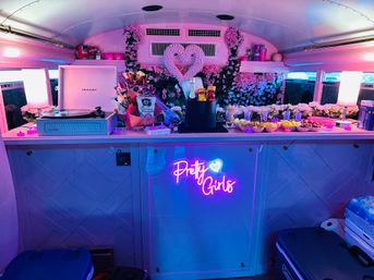 Pink-lit party bus bar interior with floral heart backdrop, vintage turntable, champagne bucket, bowls of snacks and a playful neon “Pretty Girls” sign.