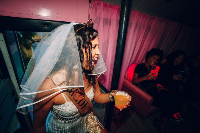 Bachelorette party scene: laughing bride-to-be in a veil and "Bride" sash holding a cocktail inside a pink-decorated party bus, friends on a couch taking photos.