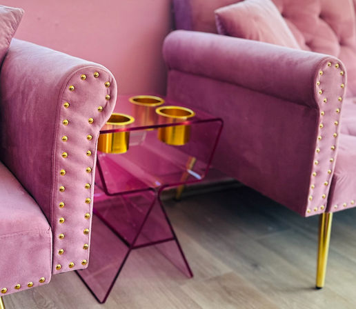 Glam pink velvet armchairs with gold studs beside a translucent pink acrylic side table holding gold cup holders on a light wood floor.