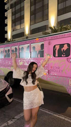 Woman in sparkly two-piece dancing at night in front of a pink party trolley with champagne-themed graphics parked on a downtown city street