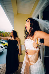 Excited bride-to-be wearing a veil and glitter sash on a sunny yellow front porch, cheering with bracelets and a smartwatch while a friend in a black dress points toward the driveway and palm trees.