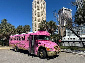 Bright pink party bus with gold trim parked by palm trees along the Tampa waterfront, downtown skyline and round high-rise under a clear blue sky