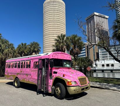 Bright pink party bus with gold trim parked by palm trees along the Tampa waterfront, downtown skyline and round high-rise under a clear blue sky