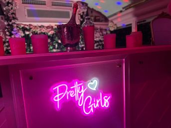 Bright pink neon "Pretty Girls" sign on a bar counter at a party lounge, with a champagne bottle in an ice bucket, pink candles, floral backdrop and colorful disco lights