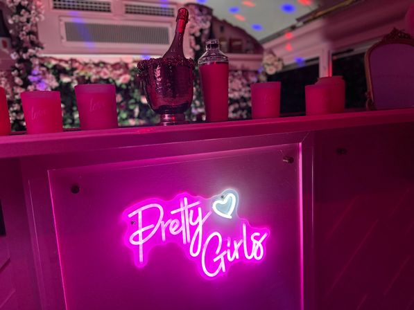 Bright pink neon "Pretty Girls" sign on a bar counter at a party lounge, with a champagne bottle in an ice bucket, pink candles, floral backdrop and colorful disco lights