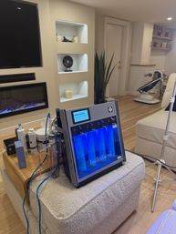 Blue-lit Hydrafacial skincare machine with clear tubing and product vials on an ottoman in a cozy home-treatment area with built-in shelves, electric fireplace, potted plant and baby rocker in the background.