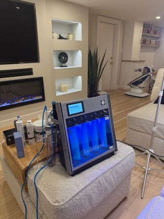 Blue-lit Hydrafacial skincare machine with clear tubing and product vials on an ottoman in a cozy home-treatment area with built-in shelves, electric fireplace, potted plant and baby rocker in the background.