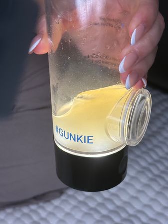 Close-up of a clear fitness shaker labeled #GUNKIE with yellow supplement residue swirling inside, held by a hand with white French-manicured nails.