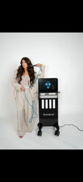 Stylish woman in a striped suit holding a Hydrafacial handpiece beside a black Hydrafacial machine in a bright skincare studio — modern facial treatment setup.