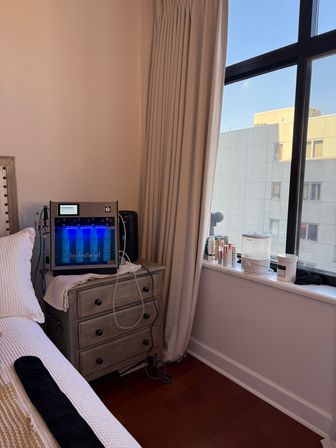 Sunlit bedroom corner with a wooden nightstand holding a glowing blue facial treatment machine, tubing, and folded towel; windowsill with skincare bottles and a white humidifier, beige curtains and cream bedding.