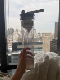 Hand holding a clear skincare treatment fluid-collection canister against a high-rise window overlooking a snowy urban skyline and city buildings
