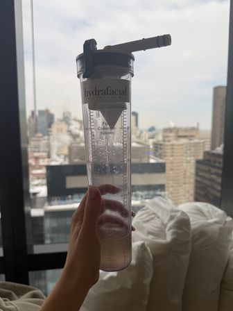 Hand holding a clear skincare treatment fluid-collection canister against a high-rise window overlooking a snowy urban skyline and city buildings