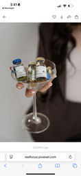 Close-up of a hand with white French manicure holding a coupe glass filled with miniature skincare ampoules and vials labeled Blue and Gold — stylish spa product display.