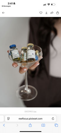 Close-up of a hand with white French manicure holding a coupe glass filled with miniature skincare ampoules and vials labeled Blue and Gold — stylish spa product display.