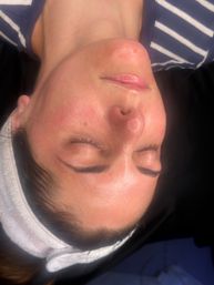 Close-up of a person lying back with eyes closed, glowing post-facial skin, freshly applied winged eyeliner and glossy lips, wearing a white headband — beauty treatment close-up.