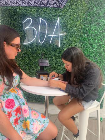 Outdoor pop-up under a neon "30A" sign on a green boxwood wall, a stylist carefully works on a customer’s wrist at a small white table — casual beach-town nail or jewelry service.