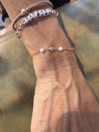 Sunlit wrist resting on an outdoor wooden table wearing layered bracelets: a delicate gold chain with two pearls and a central crystal, paired with a playful beaded bracelet of white letter cubes spelling LOVE YOU.