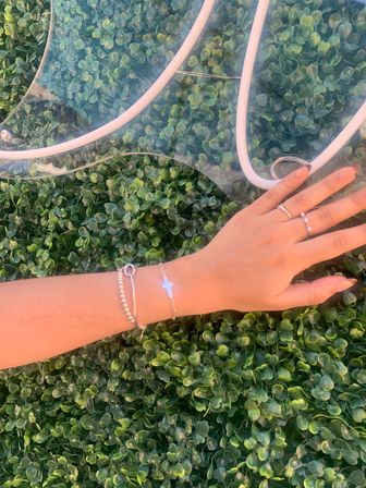 Sunlit hand with silver bracelets, a cross charm and delicate rings resting on a lush green boxwood backdrop next to a clear PVC tote