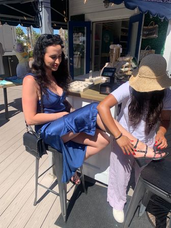 Two women at a sunny beachside boutique: one in a blue satin dress seated on a stool while another wearing a straw hat adjusts her strappy black heeled sandal by a jewelry display on a boardwalk