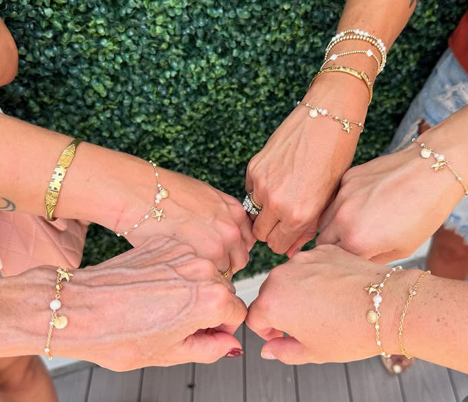 Six wrists joined in a circle showing delicate gold charm and pearl bracelets with star and shell motifs, plus rings and stacked bangles, set over a wooden deck with a green boxwood hedge backdrop