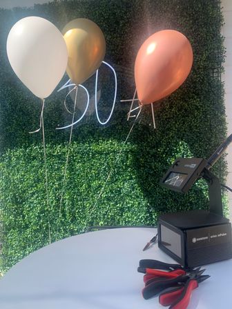 Party backdrop with three balloons — white, gold and rose-gold — floating in front of a lush green hedge and white neon '30' sign; a round white table holds a black electronic device and red-handled pliers for setup.