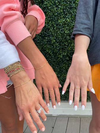 Three hands with white almond-shaped manicures wearing delicate gold chain bracelets, dainty rings and stacked beaded bracelets posed against a green hedge on an outdoor deck.