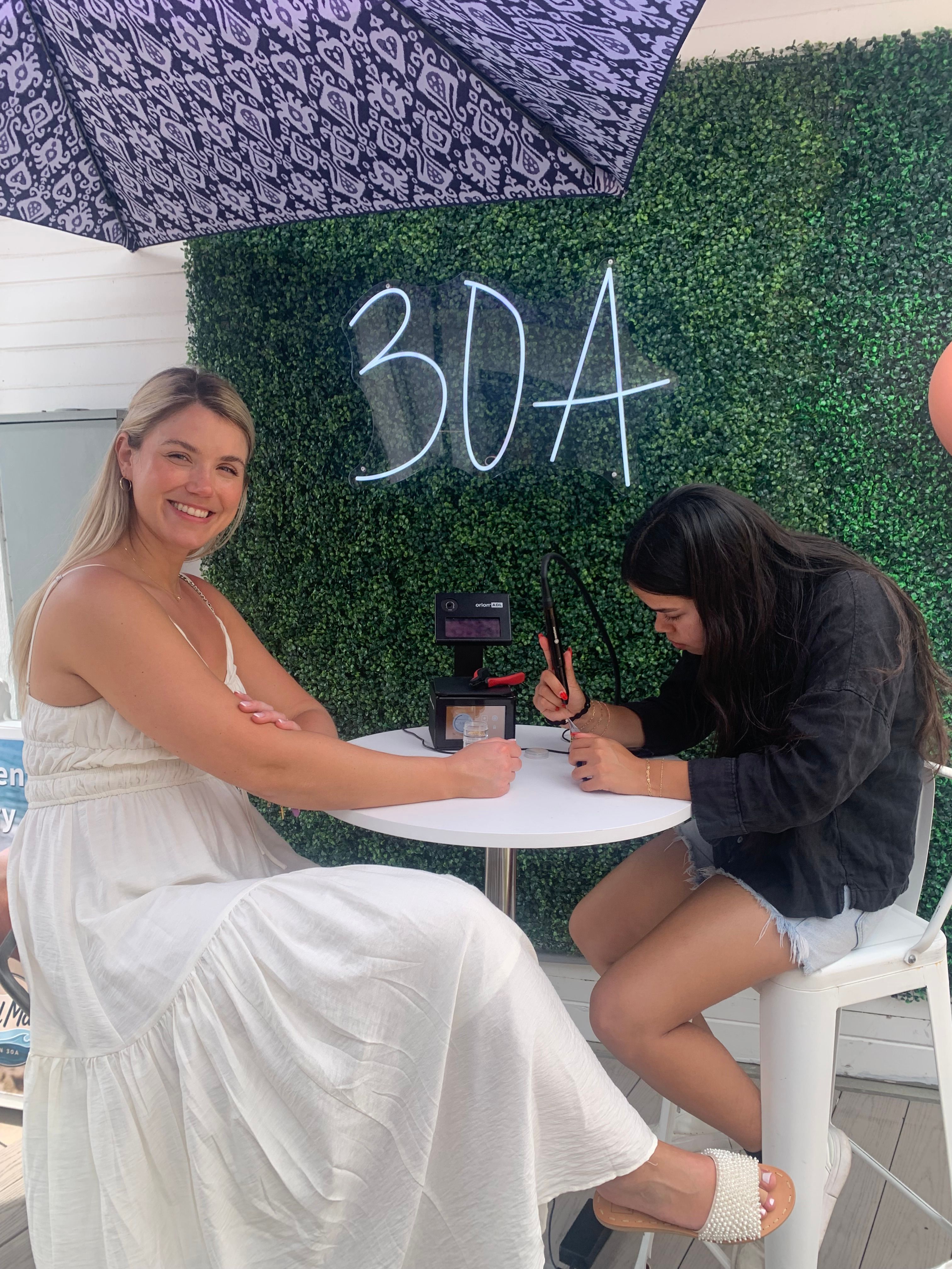 Smiling woman in a white sundress getting nail art from an artist at a small outdoor patio table under a patterned umbrella, with a green boxwood wall and neon "30A" sign behind them.